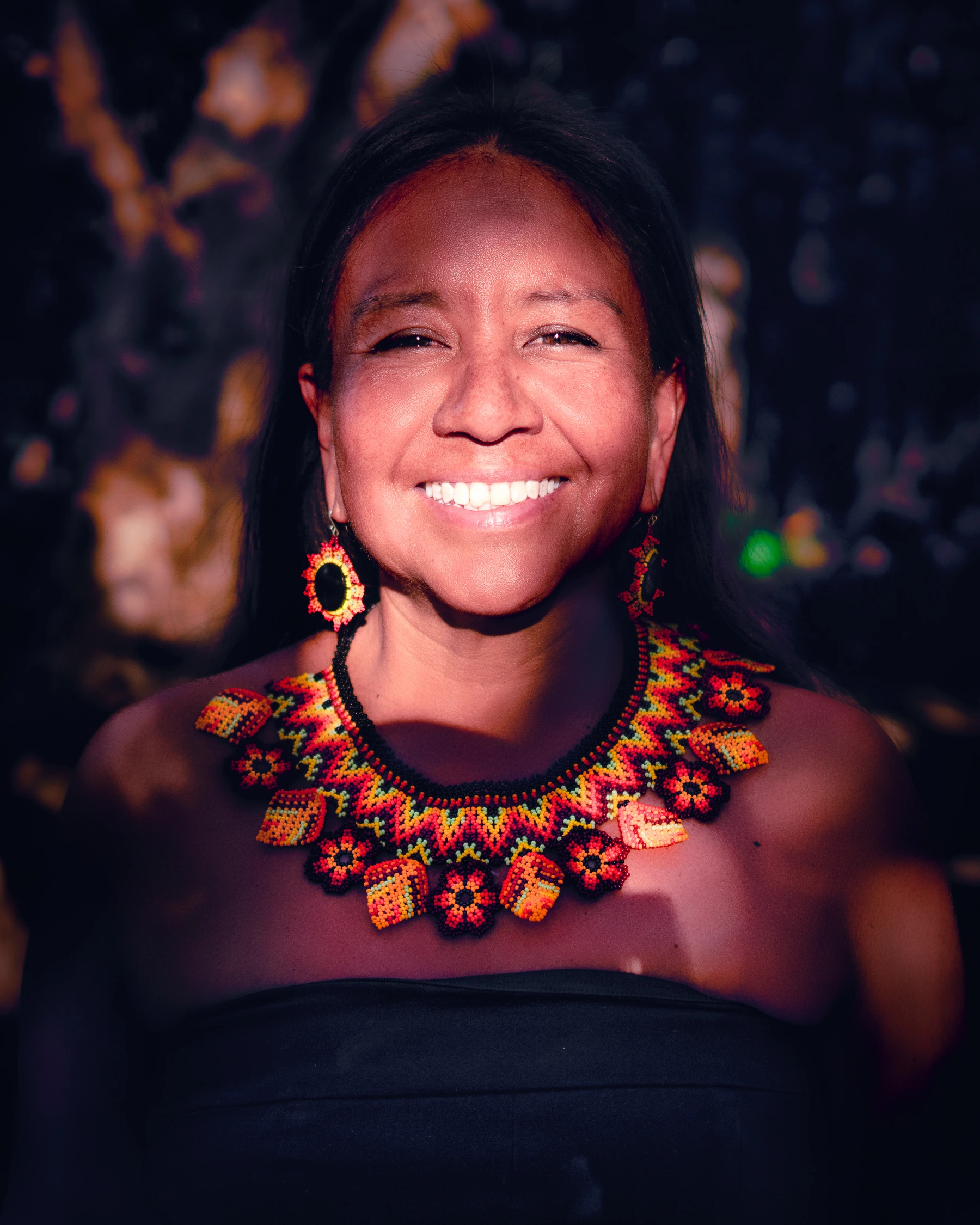 Marisela Nieves Zea (Chelita), smiling and wearing a colorful beaded necklace and earrings.