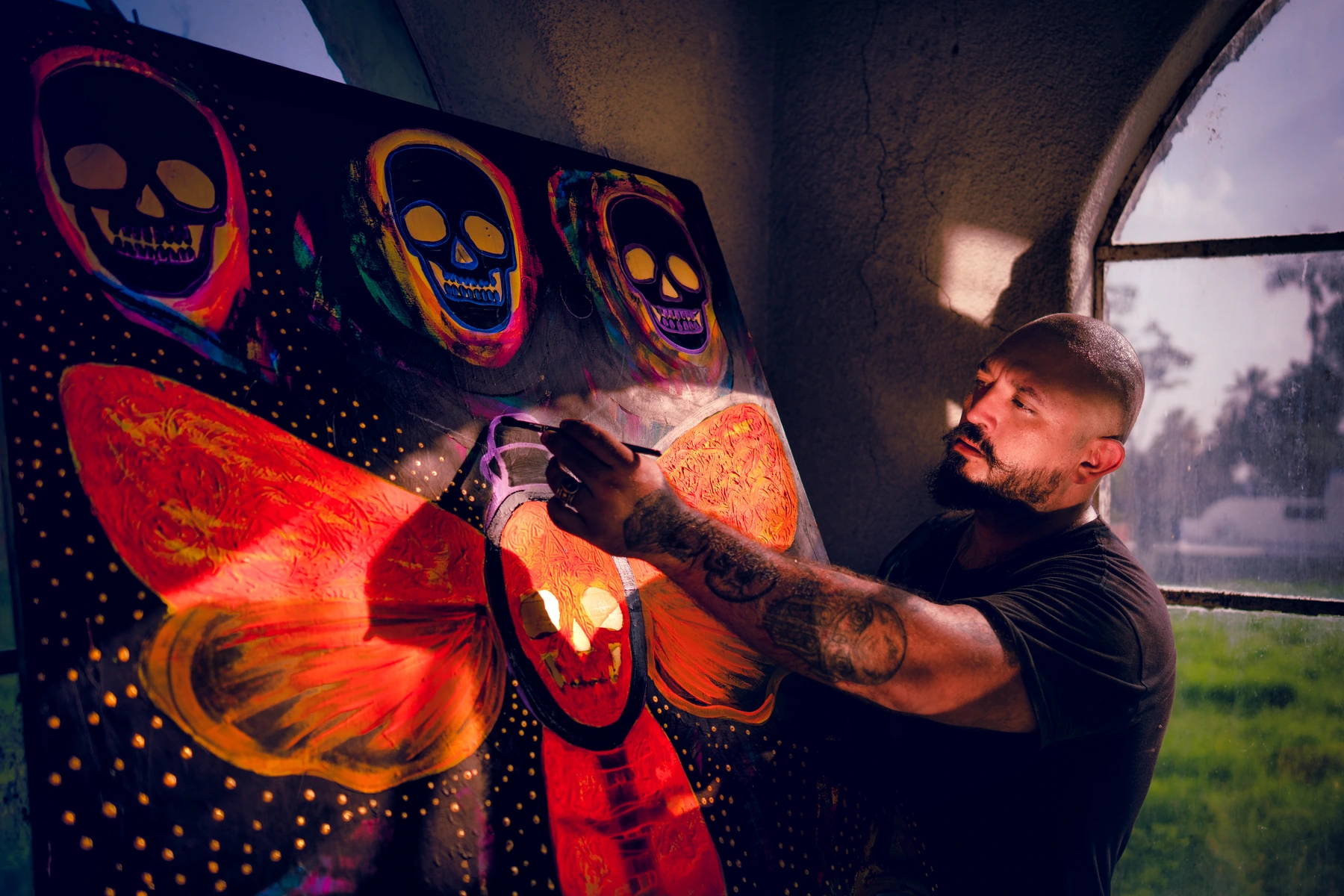 Raul Armando Prieto painting a vividly colored canvas with skull and wing motifs, next to an arched studio window.