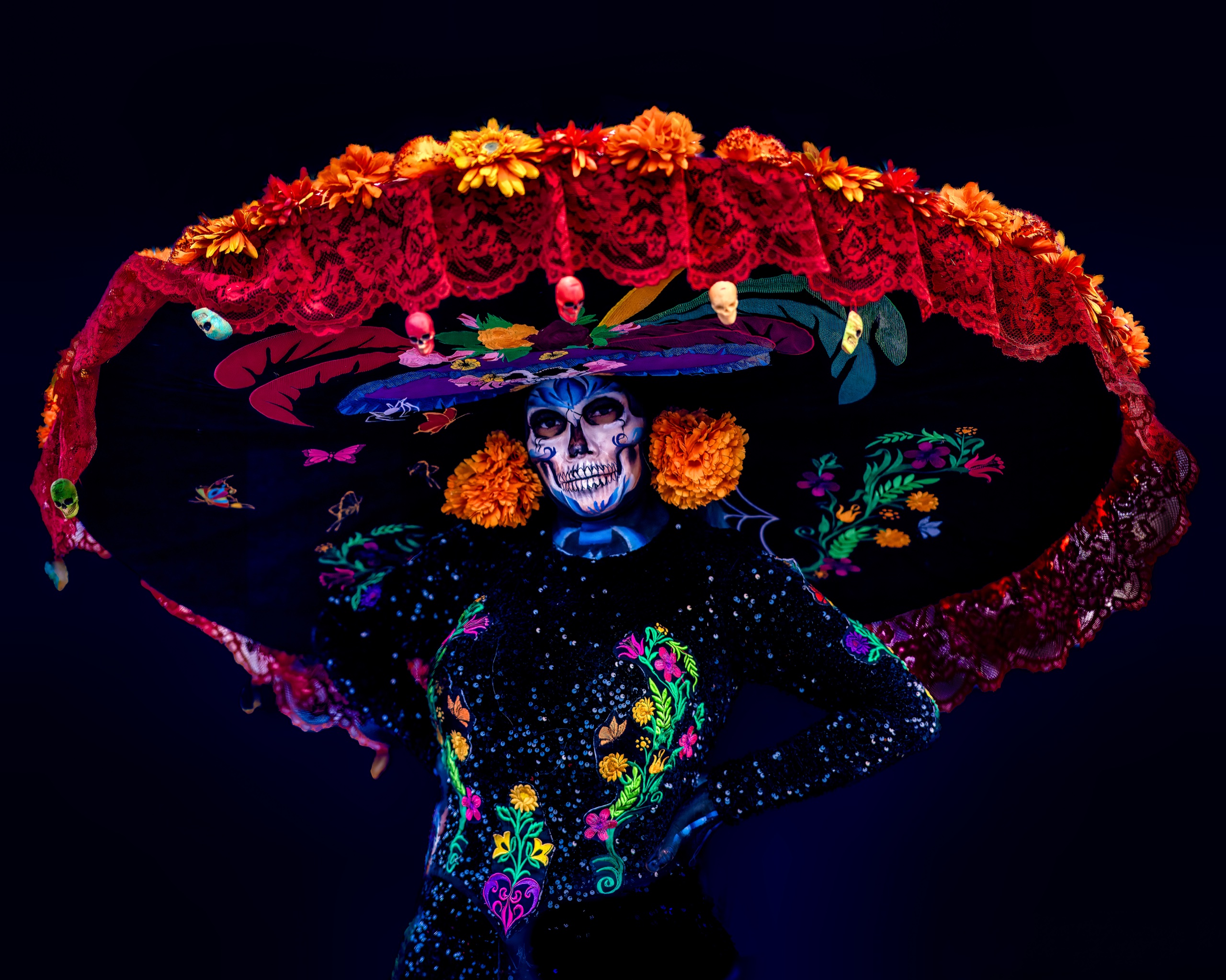 A person in elaborate Día de los Muertos Catrina makeup and a wide floral hat against a dark background.